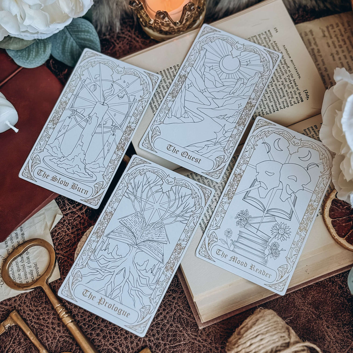 Four bookish-themed tarot cards lay diagonally across an open book, surrounded by keys, twine, a leather-bound book, a lit candle, and white flowers. The cards show tarot-inspired illustrated depictions of The Slow Burn, The Quest, The Prologue, and The Mood Reader.