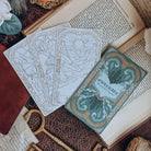 Four bookish-themed tarot cards, three face up and one face down, lie on an open book, surrounded by keys, papers, and leaves.