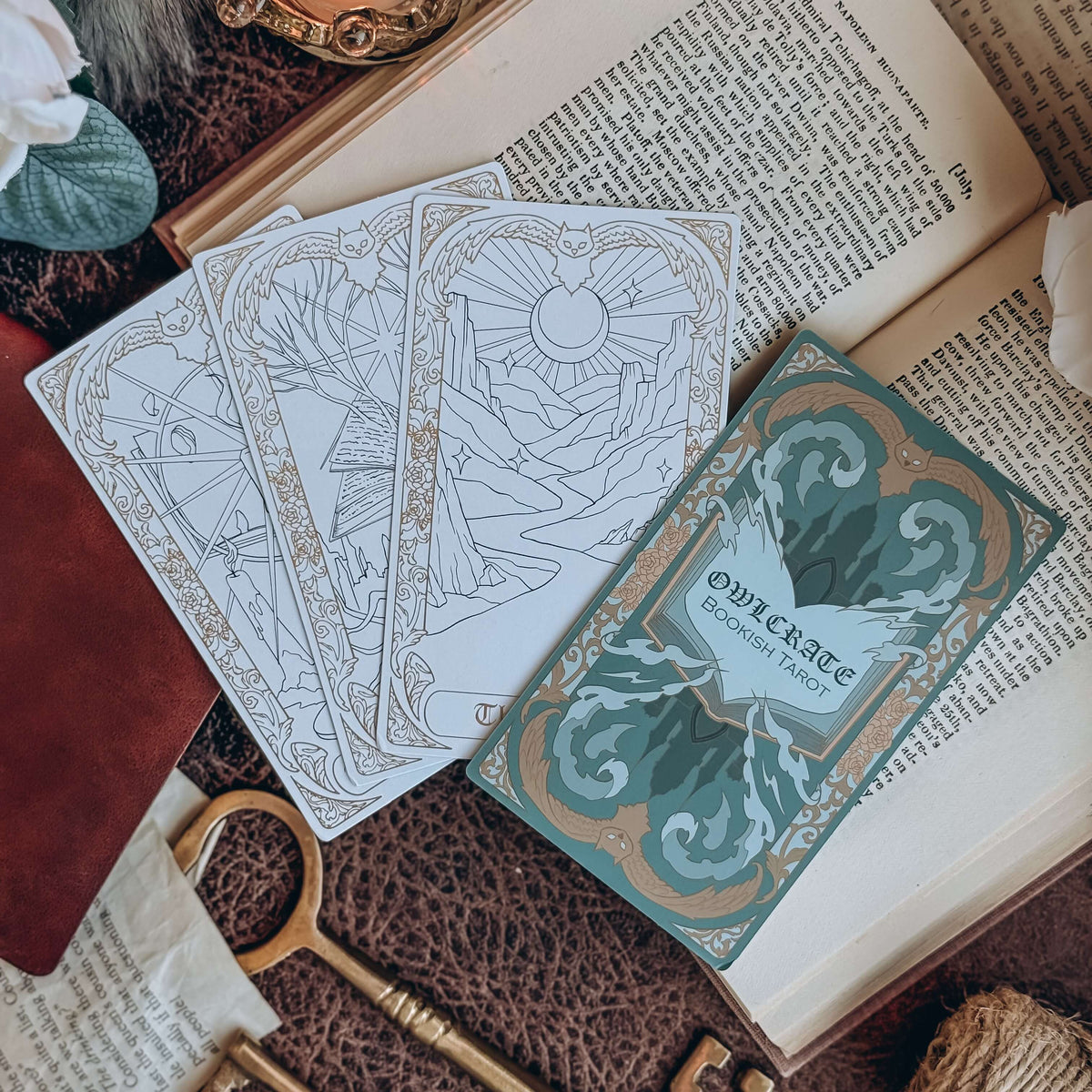 Four bookish-themed tarot cards, three face up and one face down, lie on an open book, surrounded by keys, papers, and leaves.