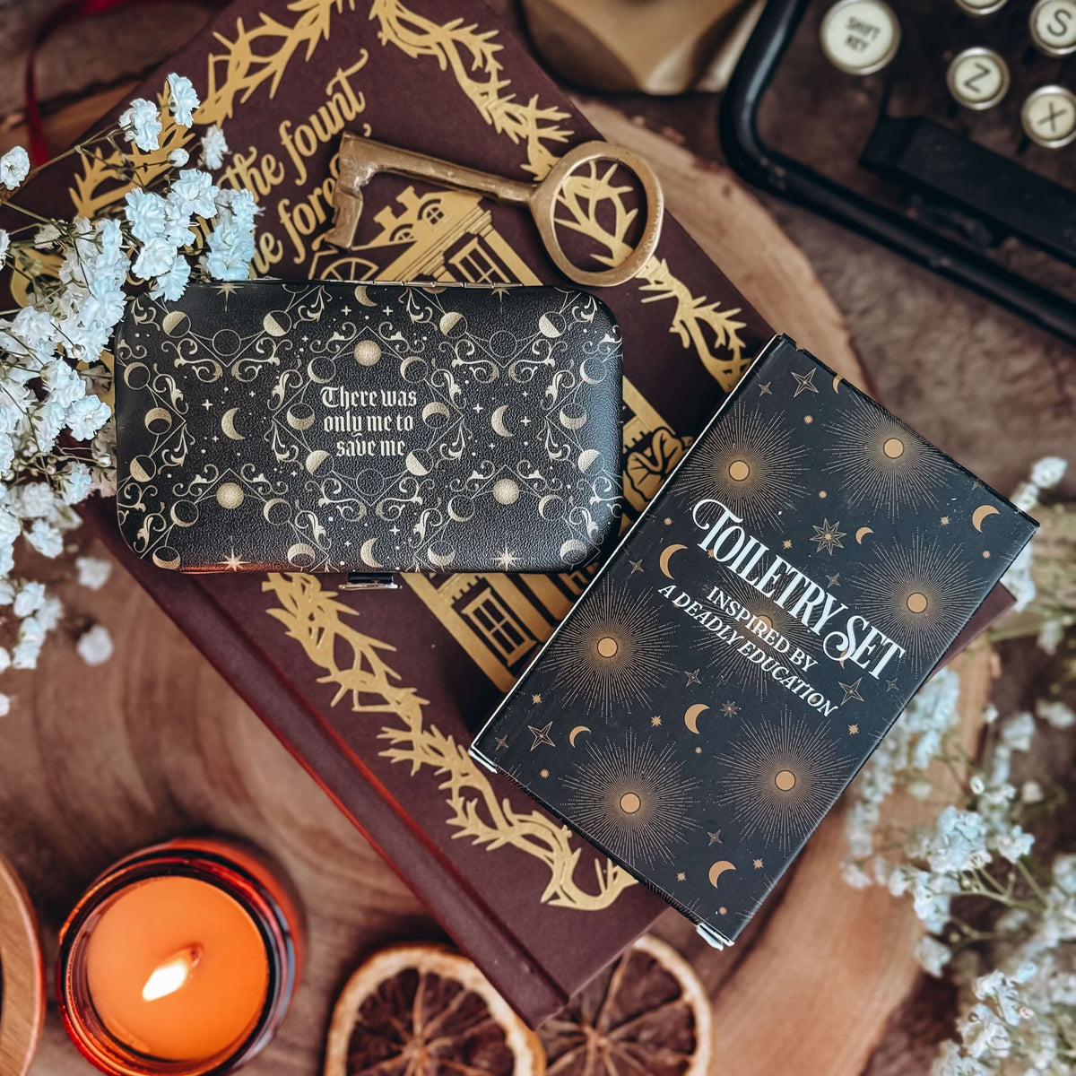 A black case and a black cardboard box of the toiletry set lies on a book with baby's breath flowers, a lit candle, and a typewriter surrounding.