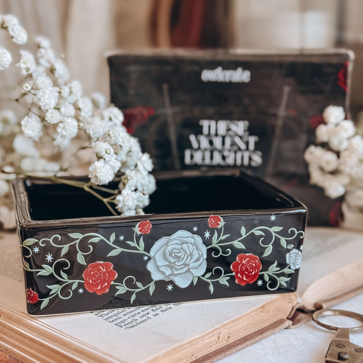 A black ceramic dish decorated with illustrated red roses, white roses, and green vines sits on an open book, stylized next to baby's breath flowers and a black, blurred branded box in the background.