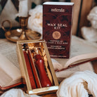 A red wax stick and a wax seal stamp sit in a gold-coloured container next to it's red kit box. Both sit on an open book with a candle stick in the background.